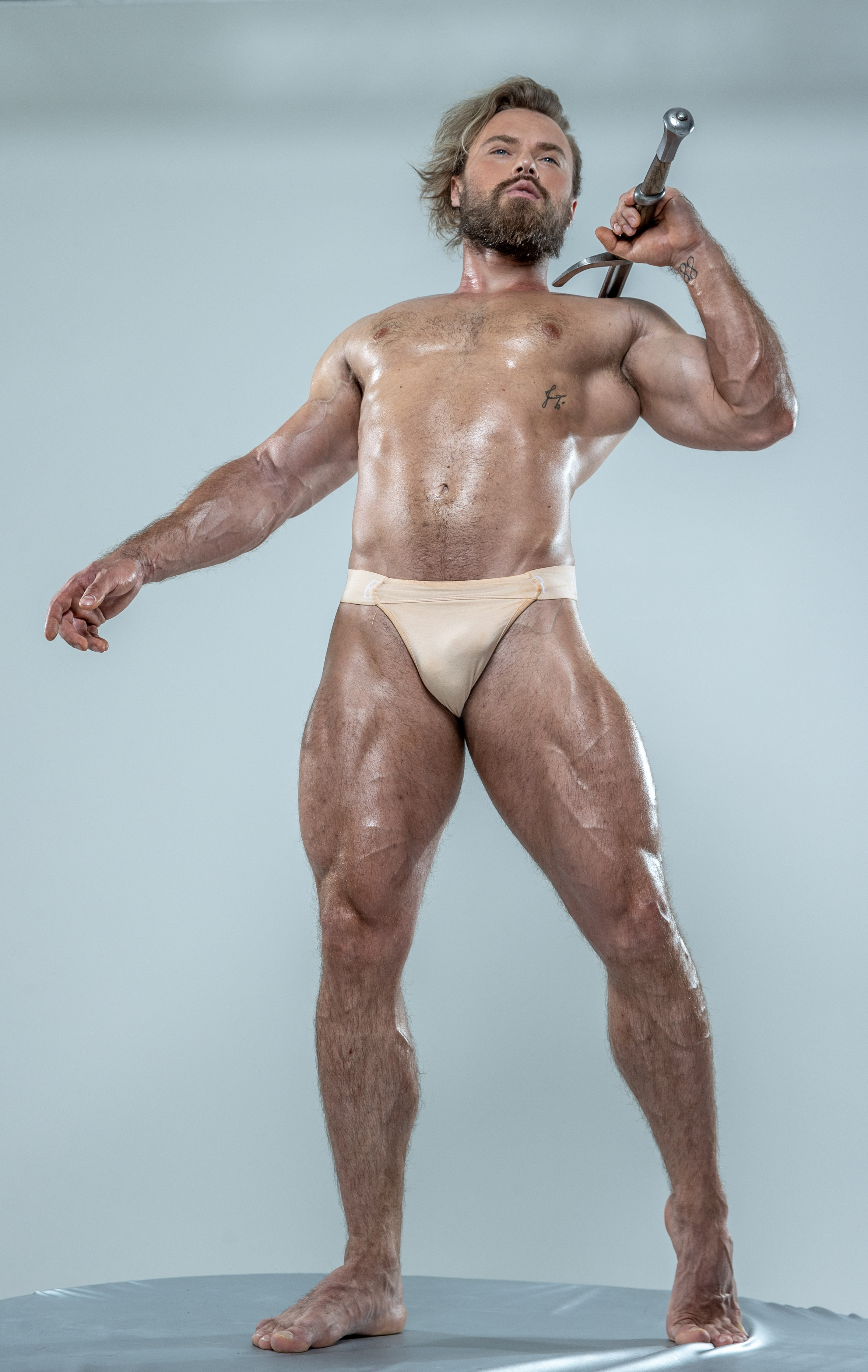 a muscular man with sweat glistening on his body stands on a small platform in a confident pose. he holds a sword over his shoulder and looks above the camera. he is wearing a  light beige speedostyle underwear. the background is a solid gray color.