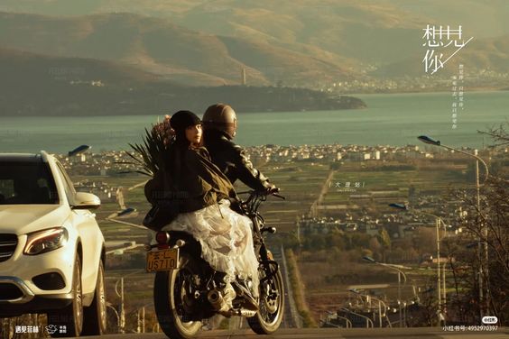 a couple is riding a motorcycle up a hillside road overlooking a scenic lake. the man is driving and the woman is riding behind him, holding a bouquet of flowers. the sun is setting in the distance, casting a warm glow over the landscape.