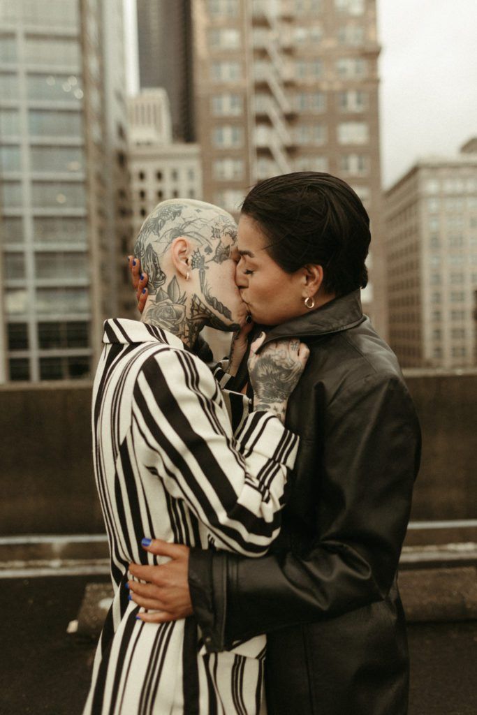 a couple standing in front of an urban cityscape, leaning in and kissing. the woman is wearing a black leather jacket, and the man is wearing a black and white striped jacket. they are both smiling, and they look very much in love. the city is a backdrop for their romantic moment. the couple are a groom and bride getting married and are embracing. they are a modern couple with a lot of tattoos, and they are very much in love.
