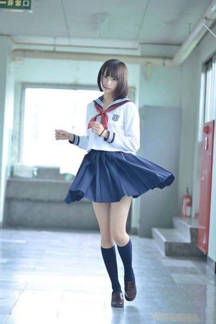 a young woman in a schoolgirl sailor uniform stands in a school hallway. she wears a white longsleeved shirt with a sailor collar trimmed in navy and a red necktie. her navy blue pleated skirt is swirling as she turns. she has knee socks in a dark color and brown leather shoes. her dark bob haircut frames her face. the corridor is bland and lit with fluorescent lights. there is a window on the left and a red fire extinguisher to the right.