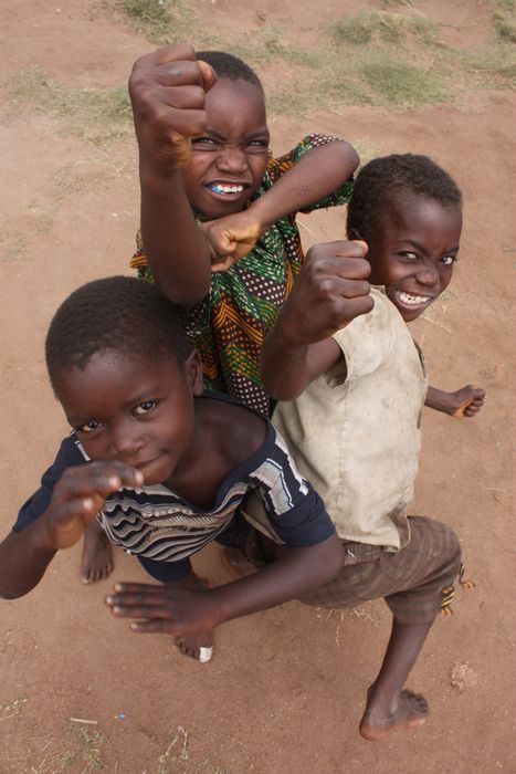 three african boys are outdoors in a red dirt environment. they are smiling and striking playful poses with their fists raised. the boys are barefoot. one boy is wearing a green and brown patterned shirt, another is wearing a white shirt, and the third is wearing a striped shirt. the image suggests a moment of fun and excitement.