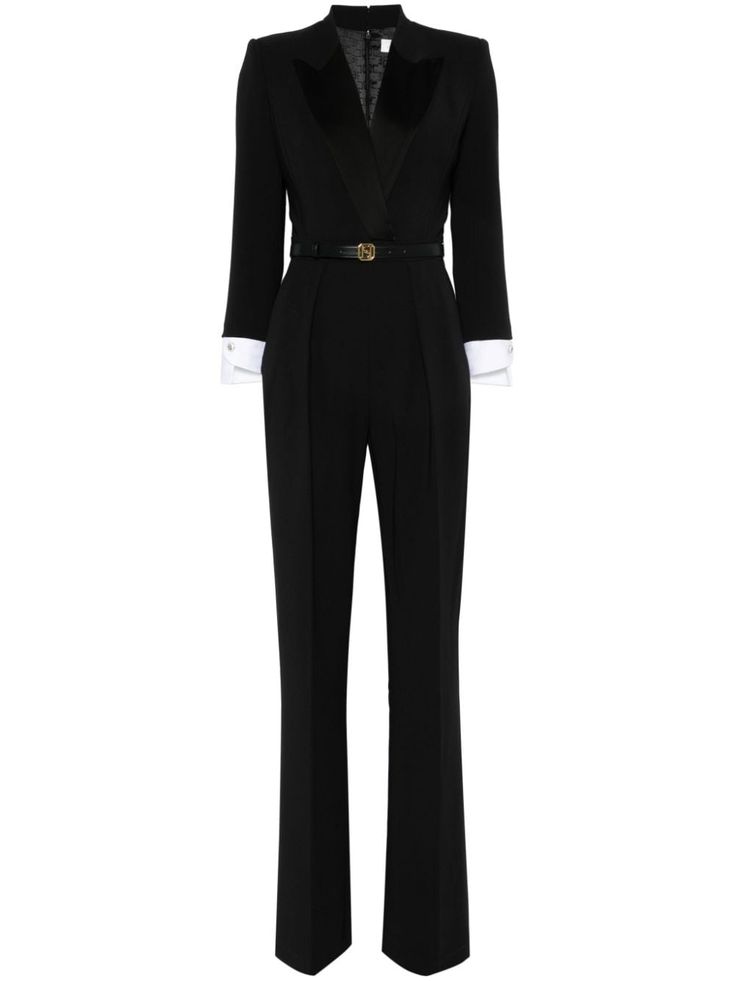 a woman is wearing a black jumpsuit with a tuxedo collar. the jumpsuit has long sleeves and a white collar. she's wearing a black belt with a gold buckle. the jumpsuit is fitted through the body and flares out at the bottom. she is standing with her arms at her sides.