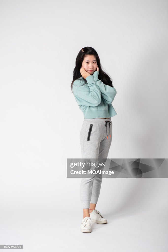 a full shot of a young asian woman wearing a light green sweatshirt and light gray sweatpants against a white background. she is standing facing the camera with her hands held up to her face. she is smiling. she is wearing white sneakers.