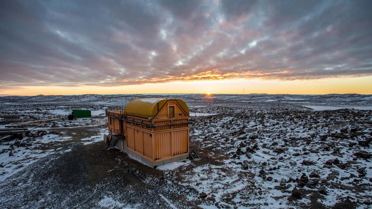 a small wooden building, likely a cabin, sits alone in a snowy, rocky landscape.  the sky is filled with colorful clouds, the sun is setting, and the horizon is far in the distance.  the scene is cold and desolate, and the building appears to be remote and isolated.