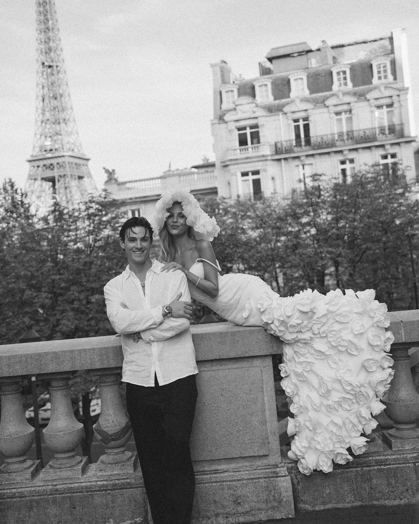 a black and white photo of a stylish couple posing in front of the eiffel tower in paris. the bride is wearing a white, floral wedding dress, and the groom is wearing a white buttondown shirt and black pants. they look happy and in love, and they are clearly enjoying their special day. the eiffel tower is a iconic symbol of romance and love, and it is the perfect backdrop for this wedding photoshoot.  the image is full of love, beauty, and joy.