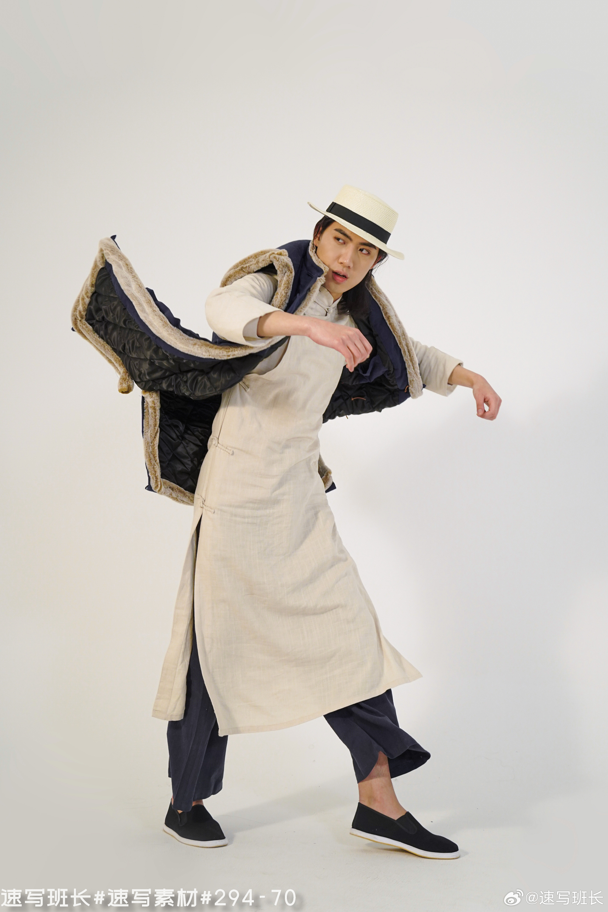a man wearing a long, lightbrown robe, a dark blue vest with fur lining, dark blue pants, black shoes, and a straw hat is striking a dynamic pose against a white background in a studio. he is looking off to the side and his arms are outstretched, with the vest's lining billowing out behind him.