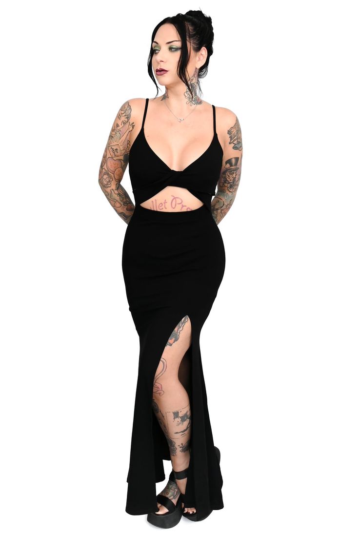 a fulllength studio shot of a tattooed woman wearing a black maxi dress. the dress is formfitting with a cutout at the waist and a high slit on one side. the woman is posing with her arms crossed and looking off to the side.  she is wearing dark platform sandals. the background is white.