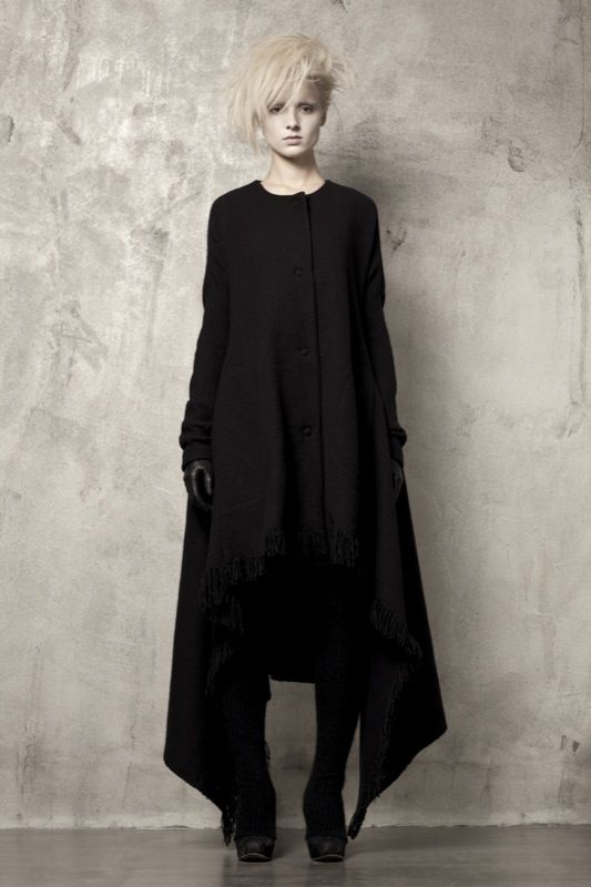 a young woman is standing against a grey wall, in a studio setting, she's wearing a black coat with fringes, and has a white, short hair, she's wearing a pair of black gloves and black shoes.  her look is chic, contemporary and monochrome.