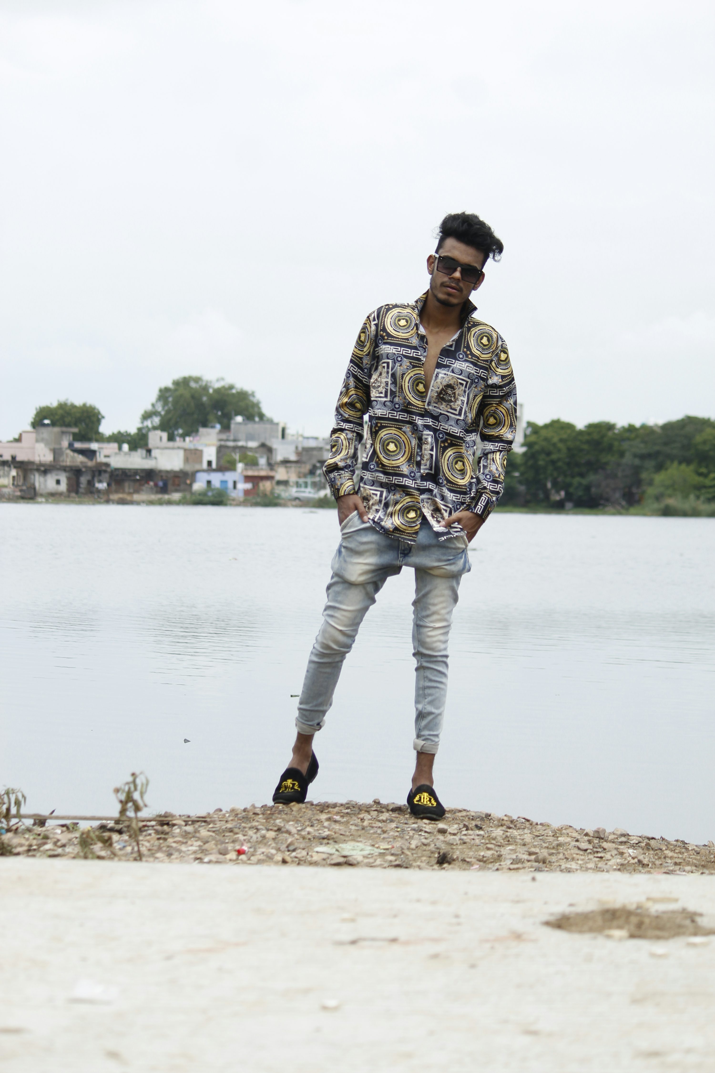 a young man, appearing to be a fashion model, stands casually by a calm lake. he is wearing a patterned shirt with a geometric design in shades of gray, black, and gold, paired with light blue jeans and black loafers. his style is relaxed yet fashionable. he's wearing sunglasses and has short, dark hair. the background features a somewhat overcast sky and a distant shoreline with buildings. the overall atmosphere is calm and serene.