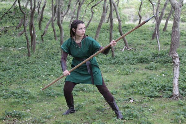 a woman dressed in an elf costume, complete with a green tunic, brown pants, and boots, holds a long spear. she is standing in a forest, adopting a fighting stance. the overall style suggests fantasy or medieval roleplaying.