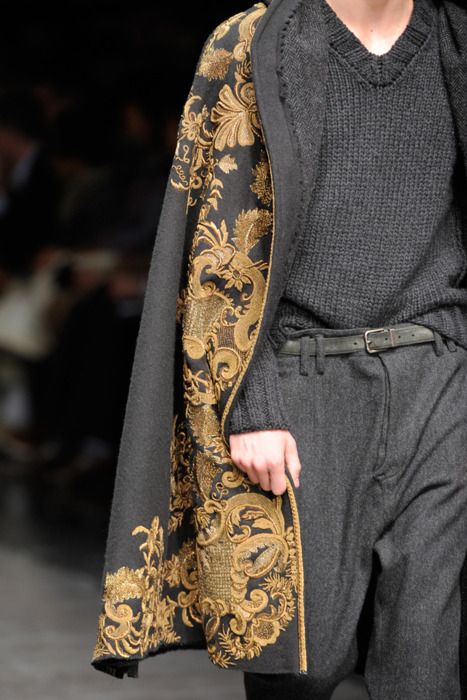 loseup shot of a male model showcasing a dark grey wool cape with intricate gold embroidery on the edges. the cape has a long, flowing design and features a rich baroquestyle pattern. underneath, the model is wearing a dark grey sweater and matching trousers with a leather belt. the image focuses on the detail and texture of the cape's embroidery, highlighting the luxurious and sophisticated nature of the garment. the background is blurred, drawing attention to the clothing. the overall style suggests an autumn/winter fashion collection.