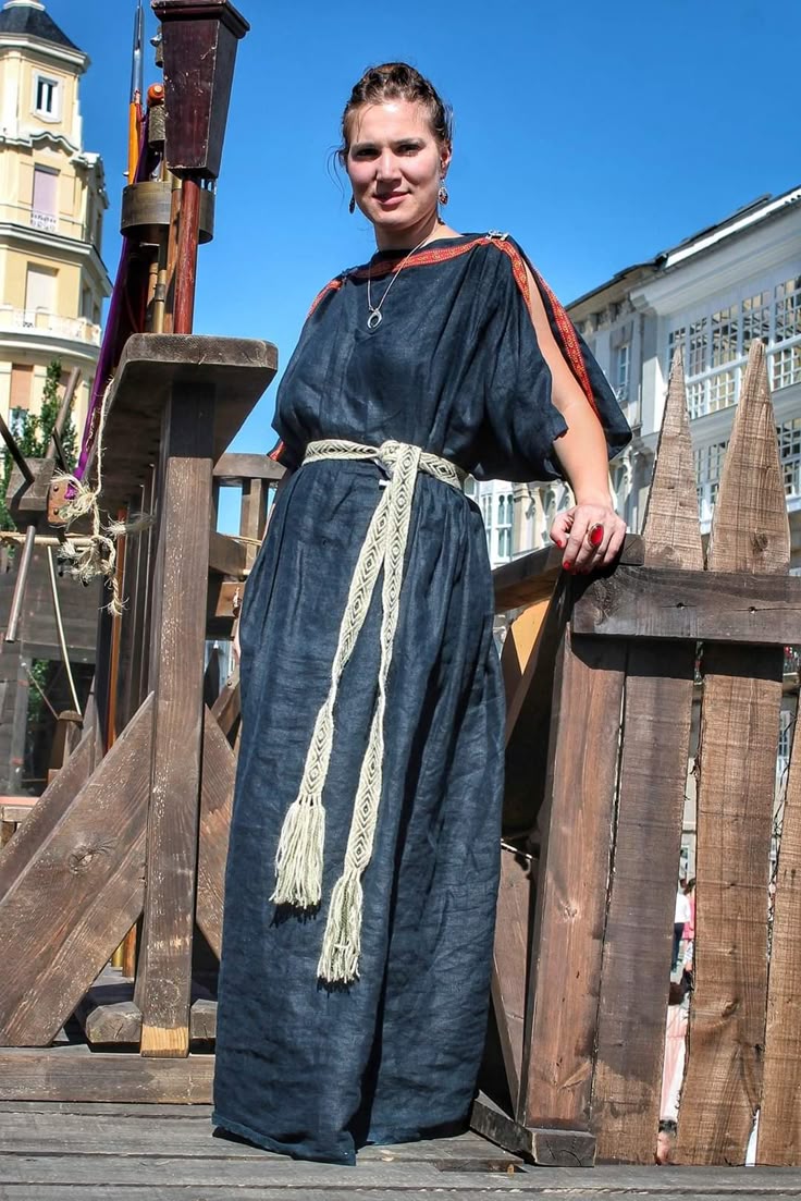 a woman wearing a dark blue historical dress with a white patterned belt stands on a wooden platform outdoors. she is also wearing jewelry, and a building is visible in the background.
