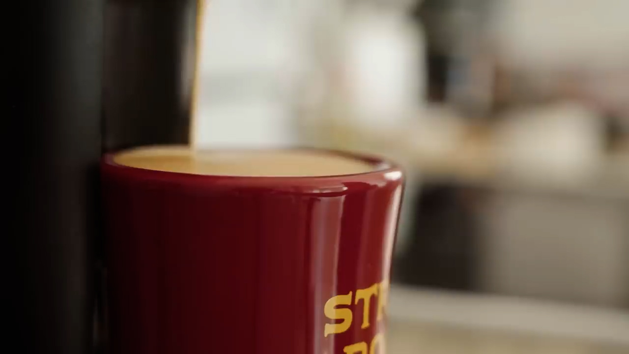 a closeup shot of a coffee maker dispensing espresso into a red "strong" mug.  the background is blurred, indicating a kitchen setting.