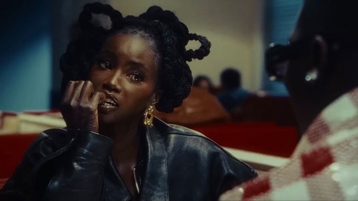 a black woman with long braids and gold earrings is sitting in a booth at a diner. she is wearing a black leather jacket and looking at the camera. a man wearing a red and white checkered shirt is in the background.