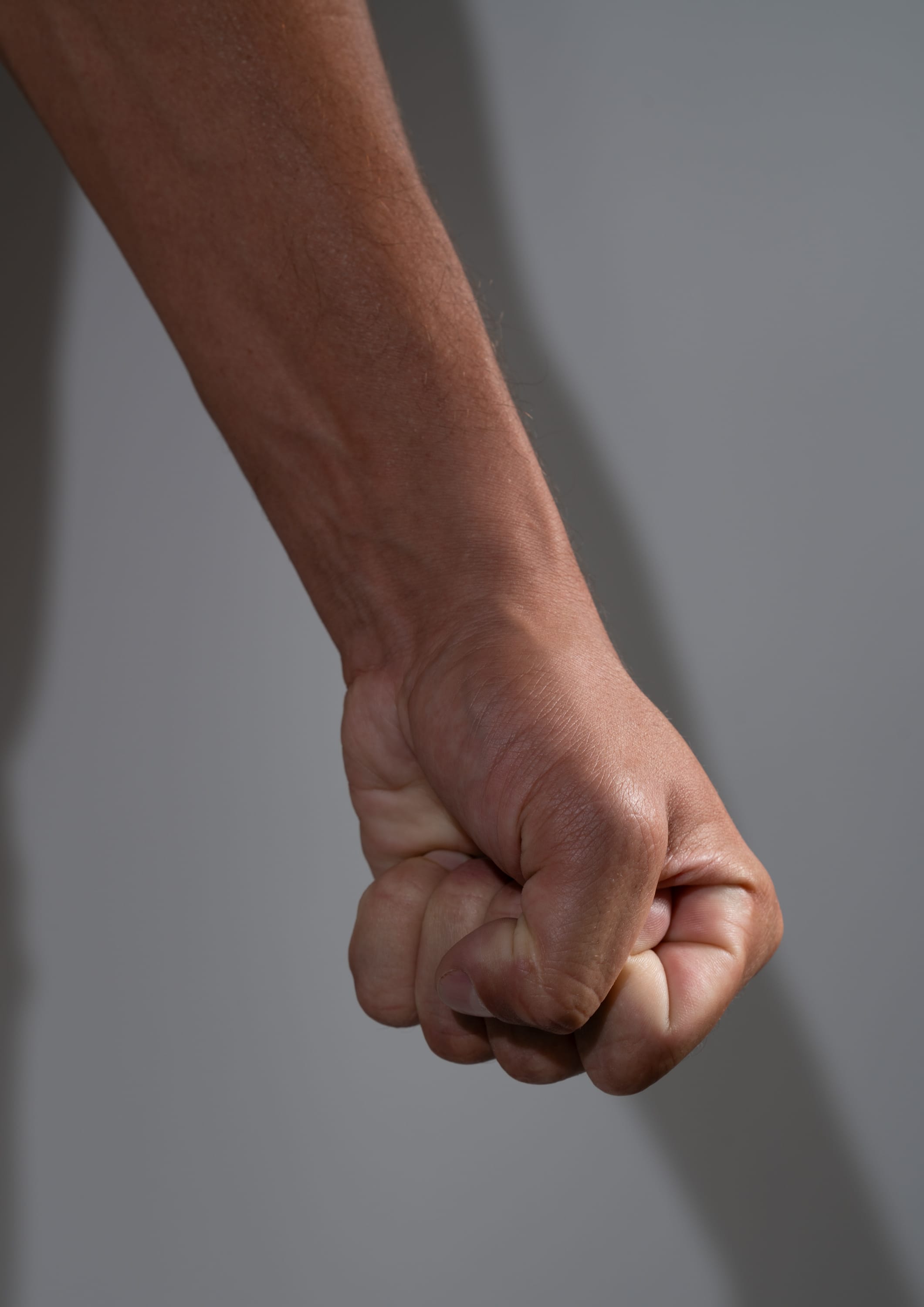 a closeup image of a clenched fist of a person with brown skin against a grey background. the fist is illuminated by studio lighting, casting a shadow on the background.