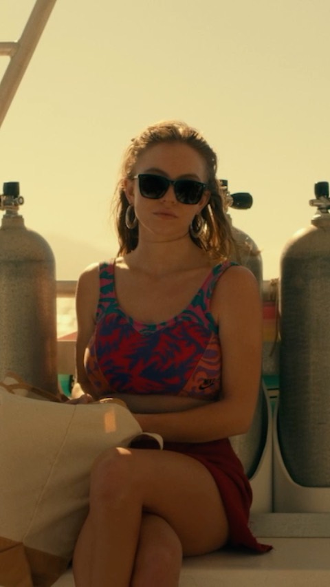 a young woman with blonde hair wearing sunglasses and a colorful tank top and red skirt sits on a boat. she has scuba diving tanks behind her, suggesting she is on a dive boat. the scene is bright and sunny, depicting a summer day at the ocean or sea.  she appears relaxed but with a somewhat serious expression.  the overall atmosphere is casual and suggests a holiday or vacation.