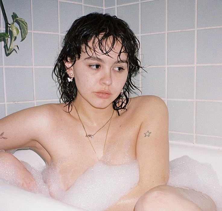 a woman is in a bathtub filled with bubbles. she has short, wet hair, a gold necklace, a star tattoo on her arm, and butterfly earrings. the background shows light blue tiles.