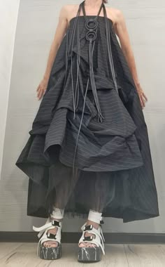 a woman in a black pinstripe dress and white platform sandals stands in a studio setting. she is wearing a black necklace and has a serious expression on her face. the dress is gathered and flowing, creating a dramatic silhouette. the shoes are chunky and have a marbled effect. she is facing the camera and her hands are to her side. the background is a simple white wall.