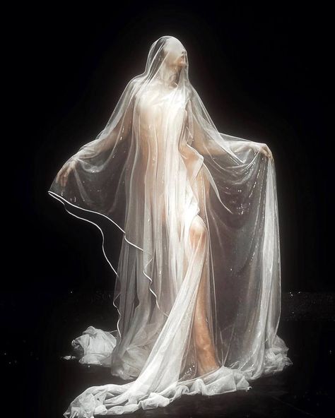 a woman in a long white dress and veil poses for a photo. the dress is sheer and billowing, and the veil is long and flowing. she is standing on a dark stage, and her silhouette is highlighted against the black background.