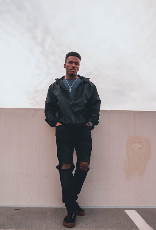 a young black man stands confidently against a pale pink wall, under a cloudy sky. he's wearing a black hooded jacket, ripped black jeans, and darkcolored sneakers with gold accents.  he has a silver cross necklace visible under his jacket.  the overall style is casual and urban.  the photo is a medium shot, showcasing his attire and demeanor.