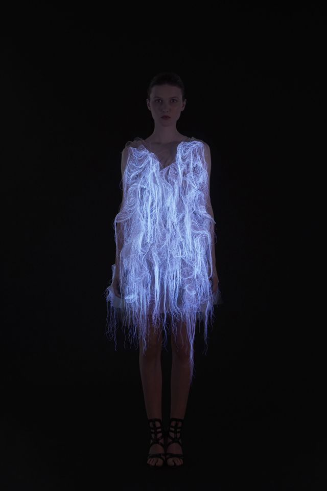 a woman wearing a glowing, fiber optic dress stands against a black background. her dress is made of white, luminous fibers and the light from the fibers casts an ethereal glow on the woman. her face is serious and she looks directly at the camera. the image is artistic and creative, and it captures the essence of innovation and experimentation in the world of fashion.