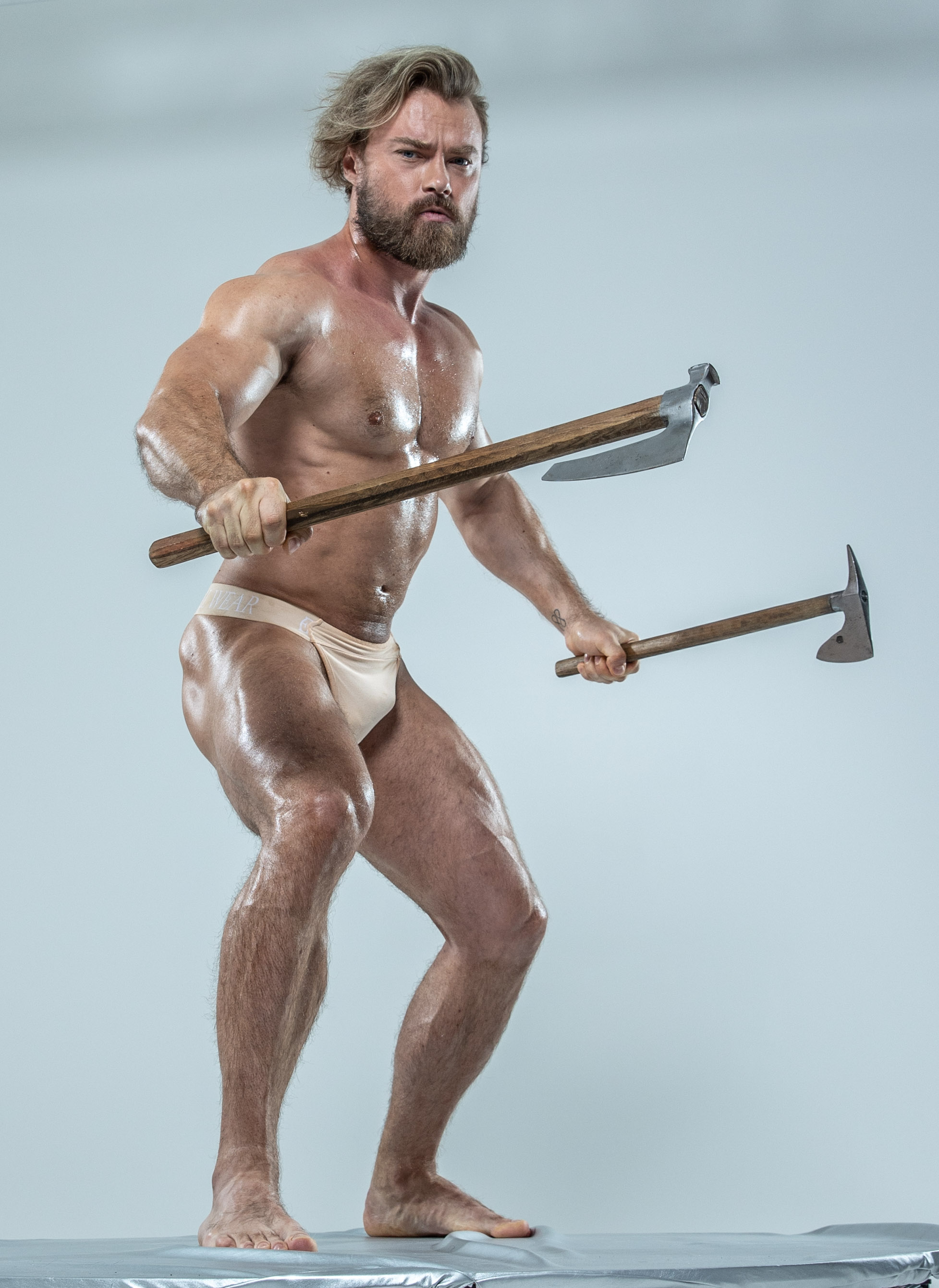 a muscular, oiled man with styled hair and beard stands on a gray platform in front of a light gray background.  he wears only white underwear and holds a large axe in each hand. his expression is serious as he looks at the camera.