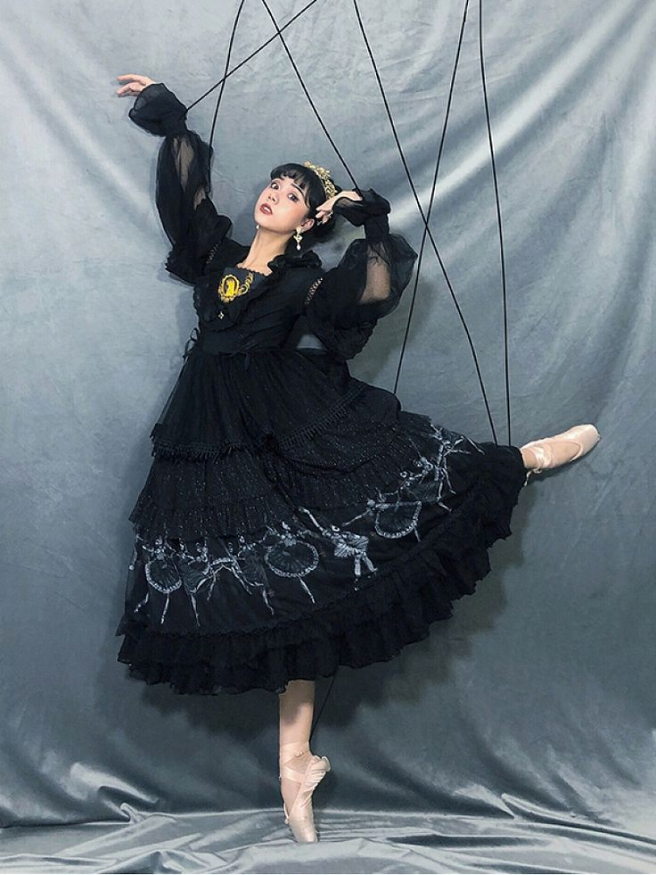 a young woman in a black dress with a ballet skirt is posing on a stage. she is wearing ballet shoes and has her leg raised in the air. the woman is looking at the camera and appears to be performing on stage.