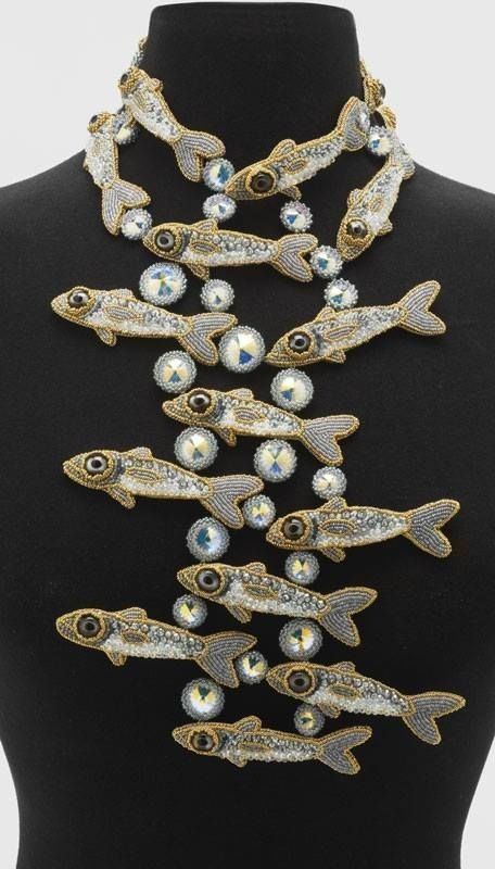 a bib necklace on a black mannequin, featuring many beaded fish and rhinestones. the fish are crafted from beads in shades of silver, gold, and white, while rhinestones are placed in between each fish. the necklace is worn on the mannequin, displaying the design and arrangement of the fish and rhinestones.