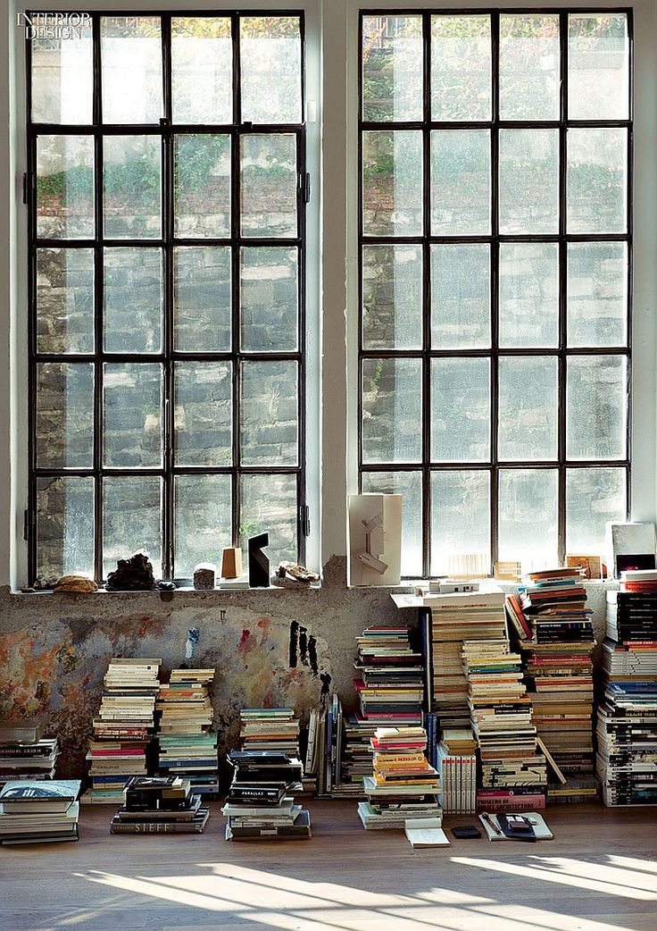 the image shows the interior of a spacious room, likely an artist's studio or loft apartment.  the room features two large, multipaned industrial windows that let in abundant natural light.  the walls are rough textured, showing signs of age and possibly paint splatters, giving the space a rustic and artistic feel.  the most striking feature is the numerous stacks of books of various sizes and colors, piled  against the wall beneath the windows.  the books are arranged in a somewhat organized, but ultimately casual way, suggesting a livedin, creative atmosphere. the overall style is vintageindustrial with a cozy, booklover's aesthetic.