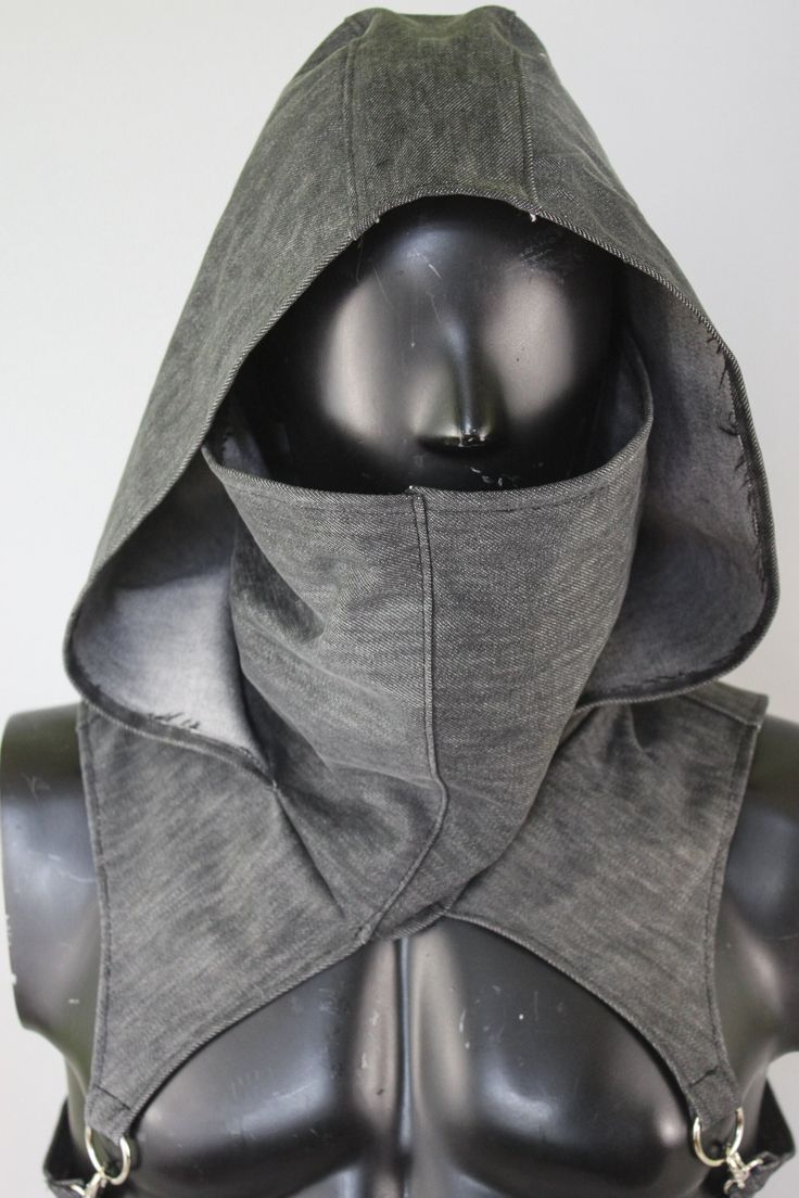 a dark gray, almost black, fabric hood and mask combination is displayed on a black mannequin. the hood has a pointed apex and drapes over the shoulders, attaching to a chest harness with metal buckles. the mask covers the lower half of the face, leaving only the eyes and forehead exposed. the overall aesthetic evokes a sense of mystery and could be associated with characters from fantasy, steampunk, or postapocalyptic genres. the lighting is even and highlights the texture of the fabric.