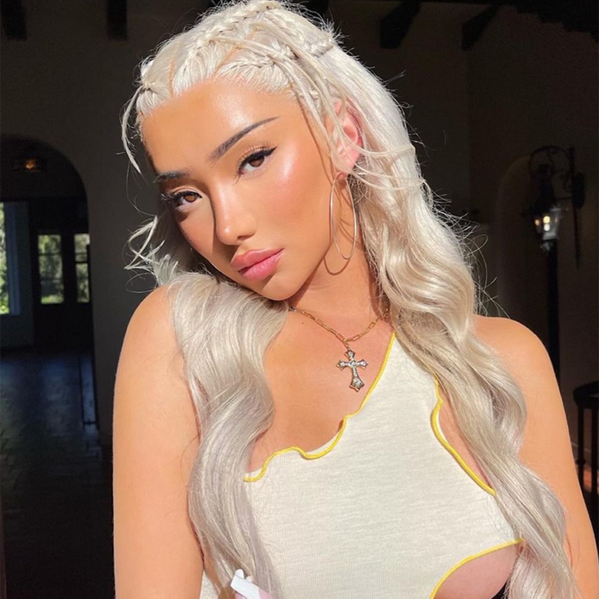 closeup portrait of a young, attractive woman with long, platinum blonde hair styled in loose waves with braids incorporated near her hairline. she's wearing gold hoop earrings, a gold cross necklace, and a creamcolored offtheshoulder top with an asymmetrical, partially cutout design.  the lighting in the image is soft and warm, with sunlight illuminating parts of her hair and face. the overall aesthetic is stylish and fashionable.