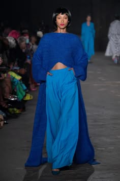 a female model walks the runway in a blue longsleeved, sweaterlike top that is tied at the waist and a long, flowing blue skirt. she has dark hair and she is striking a pose as she struts down the catwalk.