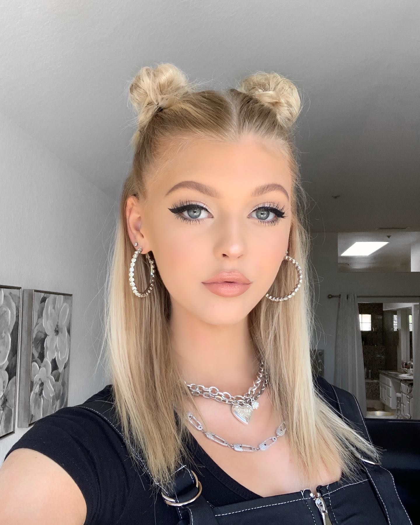 a selfie of a young woman with blonde hair styled in space buns, wearing hoop earrings, a doublelayered necklace, and a black top. she has welldefined eye makeup and is indoors with a glimpse of a hallway or room in the background.