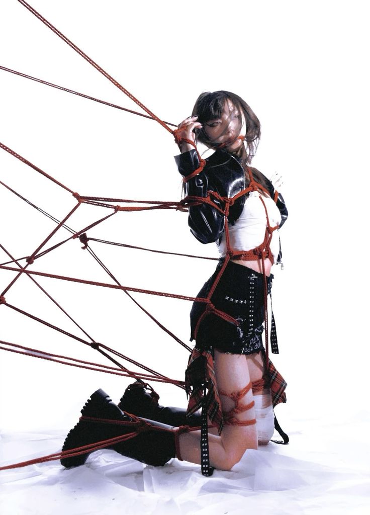 a young woman is bound with red rope in a complex shibari style.  she's kneeling on a white surface, wearing a black leather jacket, a short black skirt with studded details, and boots. the ropes are intricately woven around her body, creating a visually striking image. the background is plain white, focusing attention on the model and her bindings. the overall aesthetic is edgy and artistic, blending fashion and bondage themes.