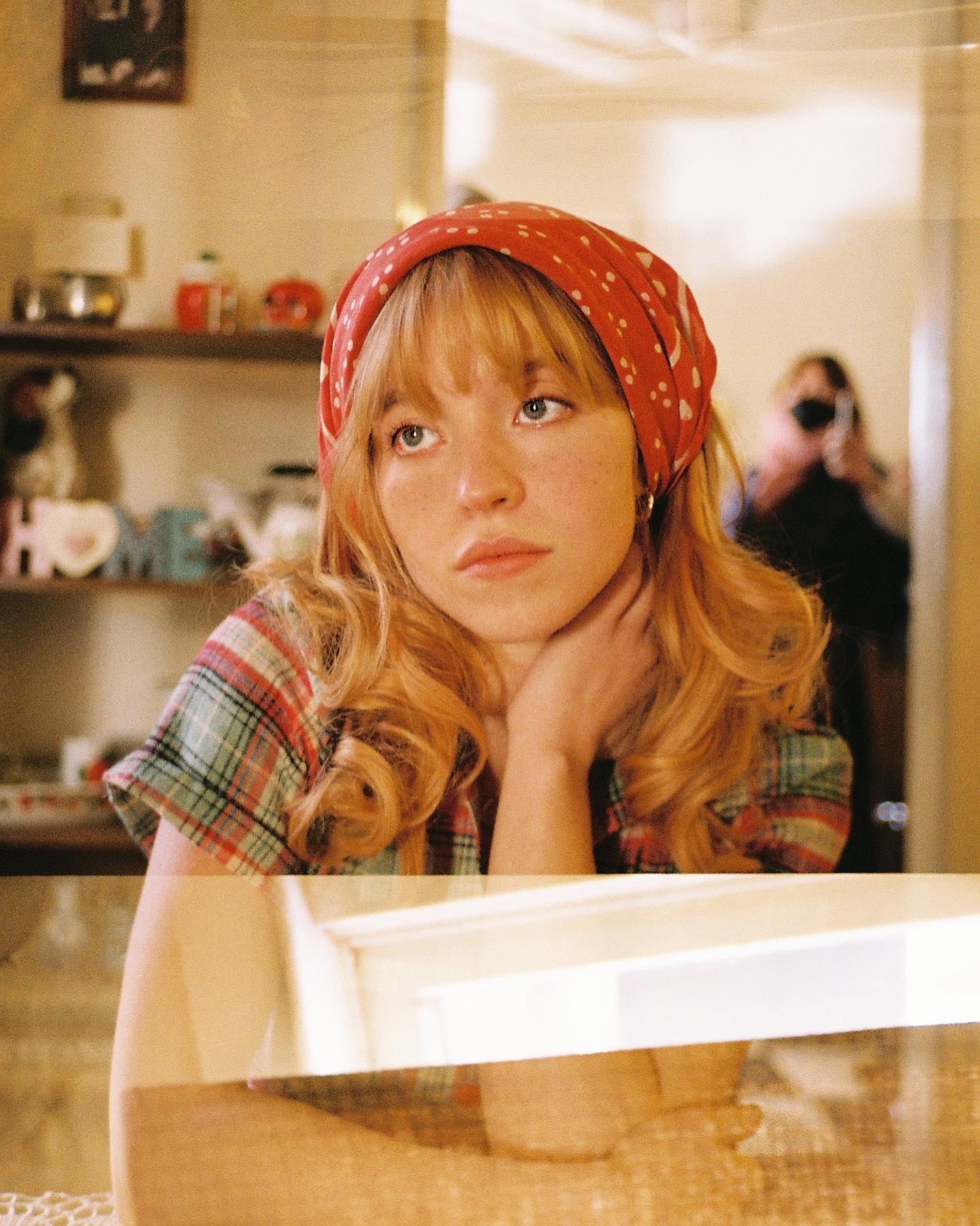 loseup portrait of a young woman with long blonde hair styled with bangs, wearing a red bandana and a plaid shirt. she is seated and looking away pensively. the setting appears to be indoors, perhaps a home environment, with a reflection of the woman visible in a mirror or glass surface. the overall aesthetic is retro or vintage, suggesting a 70s or 80s style. the image has a soft, warm tone and a slightly dreamy or nostalgic feel.  a partially visible person with a camera is in the background reflected in a glass surface.