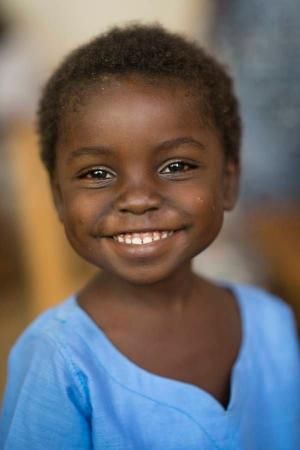 a closeup portrait of a smiling young child with dark skin. the child is wearing a blue shirt and has a happy, cheerful expression.