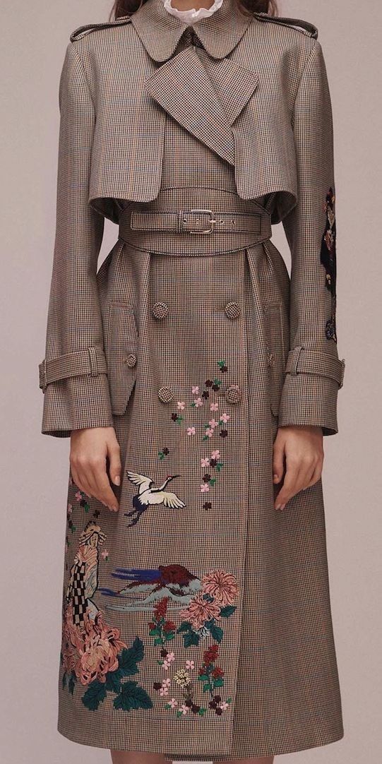 a woman is wearing a brown plaid trench coat with embroidery. the coat has a belt and a doublebreasted closure. the embroidery features flowers and birds. the woman is standing in front of a plain background.