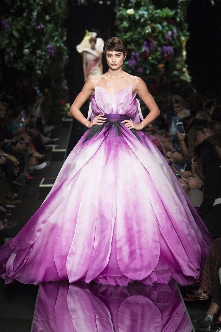 a female model walks the runway wearing a beautiful, pink, floral gown. the dress is a flowing, princesslike gown with a large,  petallike skirt. the model is wearing her hair short and has a classic, elegant look. she is striking a pose for the cameras while walking the runway.