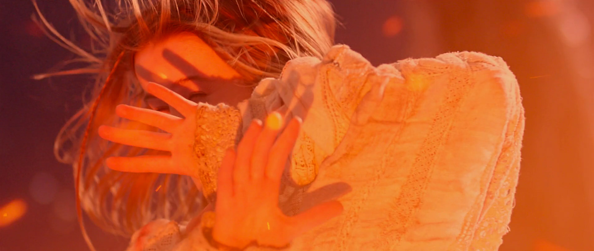 a woman with long, fiery red hair, her face hidden by a hand that is covering her eyes, stands in an orange light.  the background is blurred and indistinct, emphasizing the closeup focus on the woman's face and hand. the lighting is soft and warm, creating a dreamy, ethereal atmosphere.