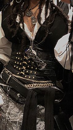 the image shows the midsection of a person dressed in a pirate costume. the costume features a black corset with gold studs and chains, a white shirt with puffy sleeves, and multiple necklaces with pendants. the person is also wearing a black leather belt with a silver buckle and a black scarf tied around their waist. the overall look is dark and edgy.