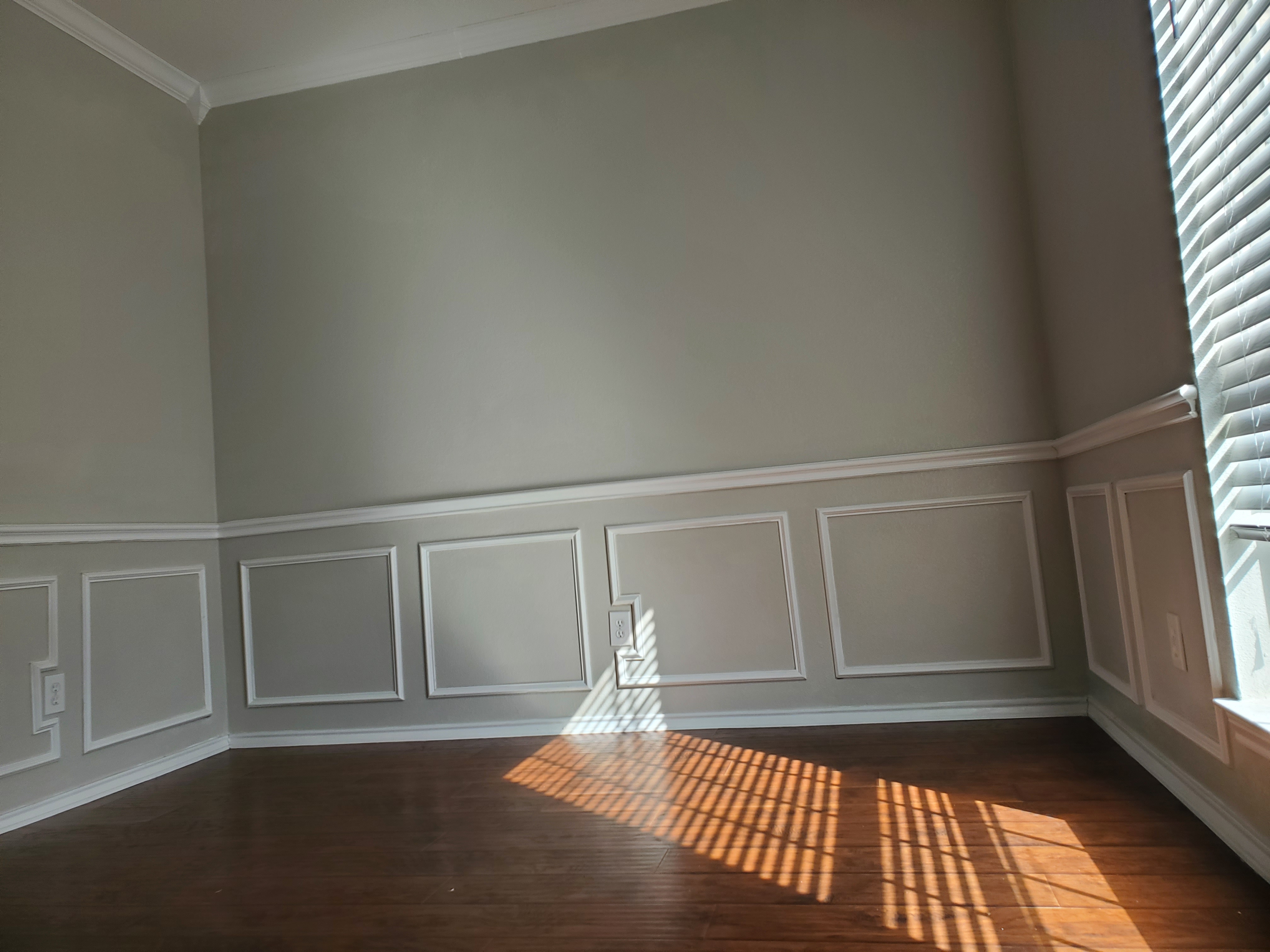 an interior shot of an empty room with hardwood floors, wall paneling with white trim, crown molding, closed window blinds with sunbeams streaming through, and a single outlet on the wall.