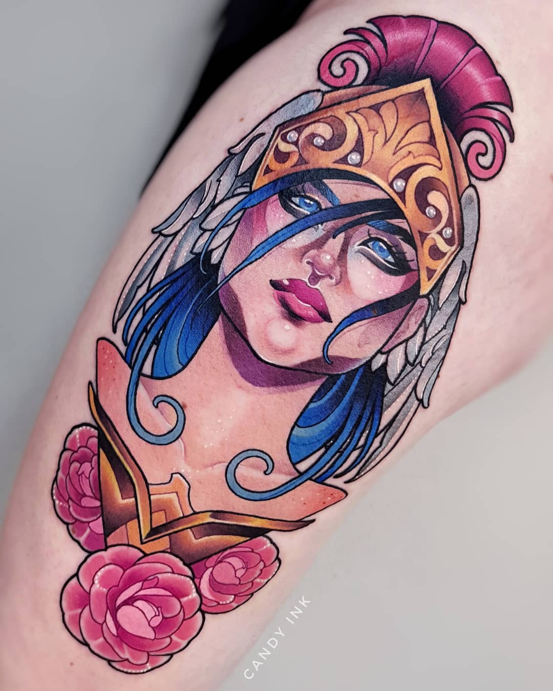 a colorful tattoo on someone's upper thigh.  the tattoo is a portrait of a woman wearing a gold greek warrior helmet with white feathers. she has blue hair, pink lips and is wearing gold armor with pink roses below it. the tattoo is very detailed and looks realistic. the background of the image is gray.