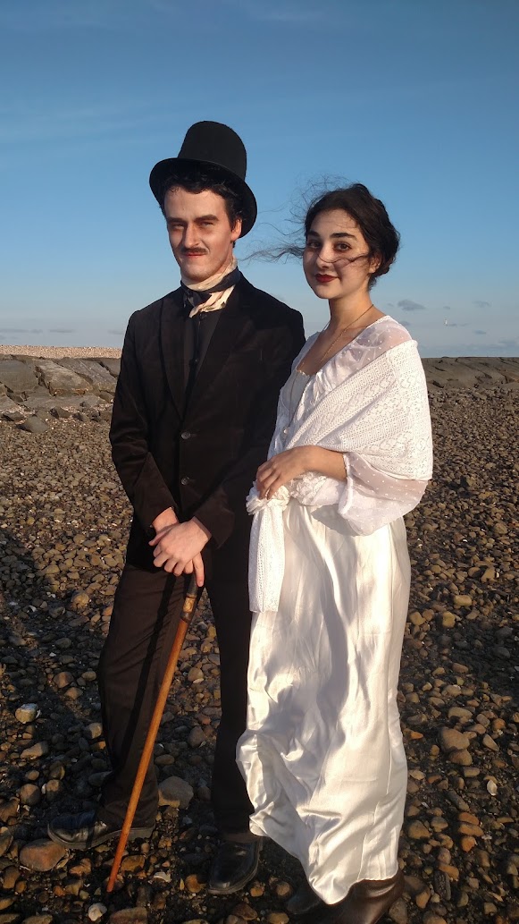 a full shot of a couple dressed in vintage 1920s1930s style clothing. the man is dressed as charlie chaplin, complete with a bowler hat, cane, and dark suit. the woman wears a flowing white dress and shawl. they are standing on a pebbled beach under a clear blue sky. the image has a historical or filmlike feel.