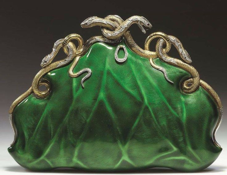 the image shows an antique ornament in the shape of a green leaf with gold and silver snakes intertwined on top.