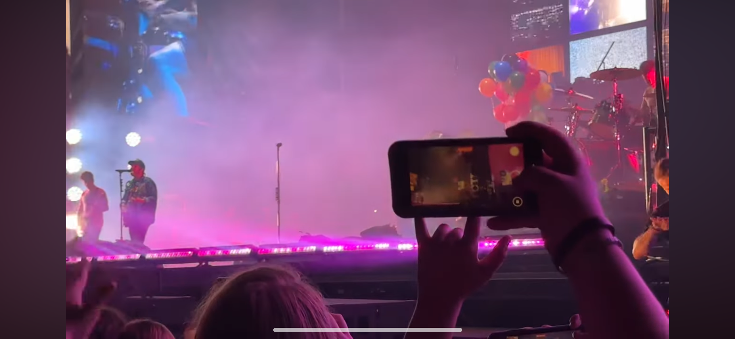 a blurry image of a concert with a person in the audience recording the stage with their cell phone. the musicians can be seen on stage playing their instruments with stage lights. the audience is blurry in the background, with the person in the foreground focused on recording the show.