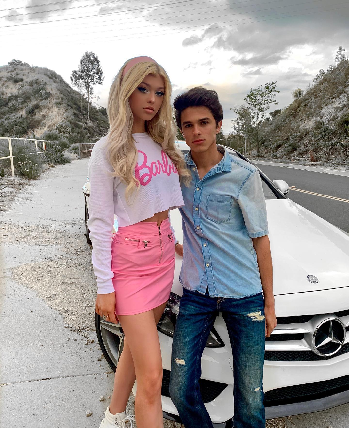 a woman with blonde hair wearing a pink skirt and a barbie crop top is standing next to a man wearing a blue denim shirt and blue jeans. they are posing in front of a white mercedesbenz car on a road with hills and trees in the background. the woman is wearing white sneakers and a pink headband.