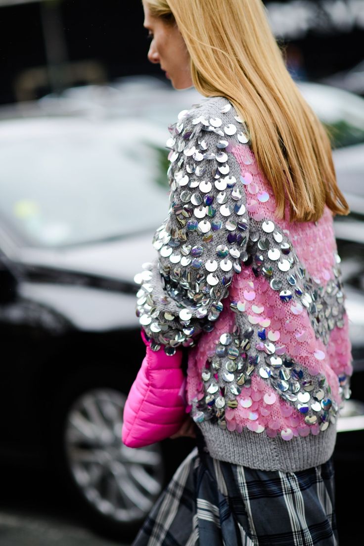 closeup view of a stylish woman wearing a grey and pink sequined sweater. the sweater has silver sequins primarily on the sleeves and shoulders, with pink sequins incorporated into the design. she's carrying a bright pink puffy handbag and wearing a plaid skirt.  her long blonde hair is straight and falls down her back. the background is blurred but shows a city street with cars.  the overall style is elegant and chic.