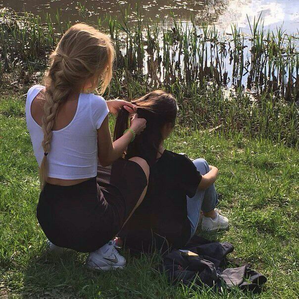 two young women are sitting on the grass by a lake. one girl is braiding the other girl's hair. the girls appear to be having a good time, smiling and enjoying the day. the background is a scenic lake with green grass and reeds.