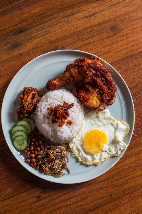 a plate of nasi lemak, a popular malaysian breakfast dish, with fried chicken, rice, egg, cucumber, peanuts, anchovies, and sambal. the dish is served on a plate with a wooden background.