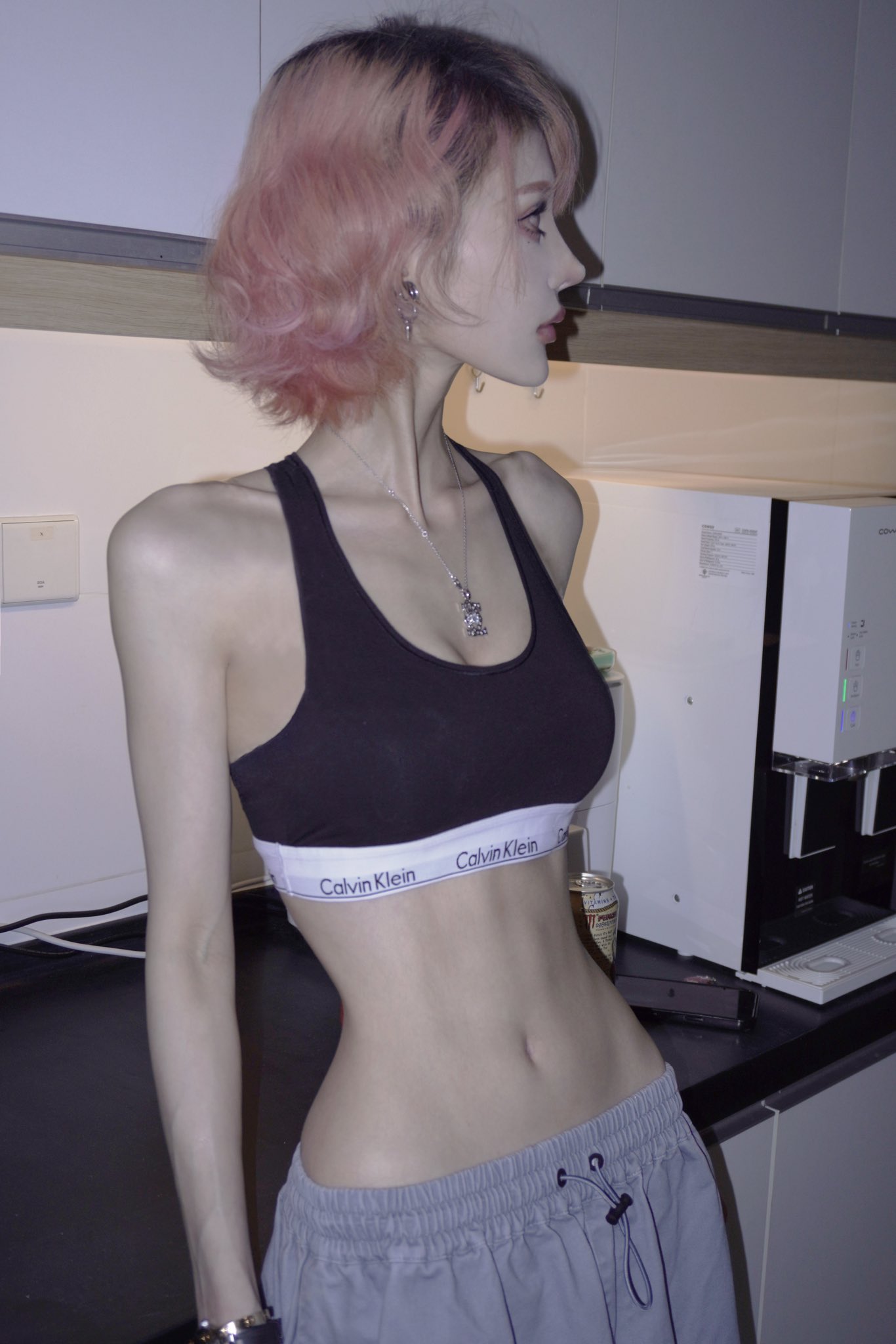 a woman with pink hair stands indoors wearing a black calvin klein sports bra and grey sweatpants. she has a necklace on and is viewed from the side. she appears very slim.